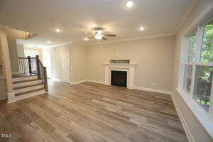 A spacious, freshly renovated living room features light hardwood floors and beige walls. There is a ceiling fan, recessed lighting, a white fireplace, and large windows. A staircase with a dark railing is visible on the left.