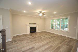 A spacious, empty living room with light-colored walls and wood flooring. It features a ceiling fan with lights, a fireplace with a mantel, and a large window that overlooks a porch with trees outside.