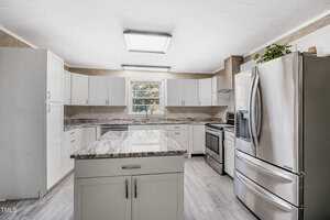 A modern kitchen with white cabinets, stainless steel appliances, and a central island with a marble countertop. Natural light enters through a window above the sink. The floor is light-colored hardwood.