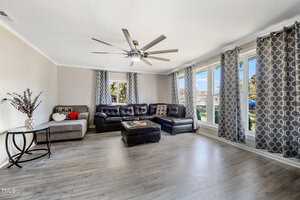 A spacious living room with a large brown sectional sofa, gray armchair, and matching ottoman. A ceiling fan is centered above. Multiple windows with geometric-patterned curtains allow natural light. A side table with a vase is on the left.