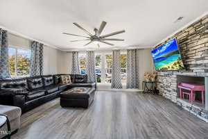 A spacious living room with a large L-shaped black leather sofa, a coffee table, and a flat-screen TV mounted above a stone fireplace. The room has wood flooring, multiple windows with patterned curtains, and a ceiling fan with multiple blades.