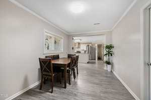 A dining room with a wooden table and six matching chairs on a light gray wood floor. A window with blinds is on the left wall. Two potted plants are in the background near the entrance to the kitchen, which has white cabinets and appliances.
