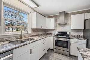 Modern kitchen with white cabinets, stainless steel appliances, and a double sink under a large window. The countertops are dark with a marbled pattern. A range hood is above the stove, and gray tile flooring complements the neutral color scheme.