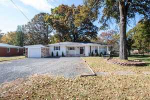 Tucked away in Butner, this charming single-story white house at 303 13th Street features a gray roof and is embraced by lush trees. A cozy front porch with chairs invites relaxation, while a single-car garage lies to the left. The driveway is speckled with leaves, and a tree in a circular brick border graces the front yard.