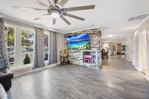 A living room with light wood flooring and a stone fireplace featuring a flat-screen TV displaying a beach scene. Large windows with patterned curtains allow natural light. A ceiling fan is in view, with a corridor leading to other rooms.
