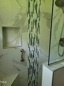 A modern shower with white and gray marble tile, featuring a vertical accent strip of smaller rectangular tiles. There's a built-in shelf niche, a metal grab bar, and a glass shower door. The showerhead is visible on the right side.