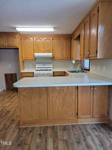 A kitchen featuring wooden cabinets and a light-colored countertop. It includes a white gas stove and an overhead range hood. The floor is finished with a wood-look material, and there is overhead lighting.
