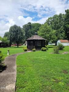 A garden scene with a circular gazebo at the center, surrounded by a well-maintained lawn. A curved concrete pathway leads to the gazebo, with trees, shrubs, and a shed in the background under a partly cloudy sky.