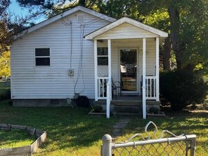 A small, single-story white house with a covered front porch and two chairs. The yard is grassy with a sectioned-off garden area. A chain-link fence is in the foreground, and trees surround the house.
