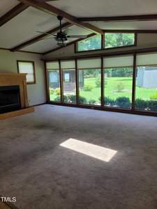 A spacious, empty living room with carpeted flooring, a wooden ceiling beam, and a ceiling fan. Large windows offer a view of a lush green backyard and garden. There's a fireplace on the left wall. Bright sunlight illuminates the room.