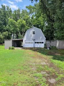 A gray, barn-style garage with two white doors is surrounded by a wooden fence. The building has a small overhang on the left side with equipment underneath. The background features tall green trees under a partially cloudy sky.