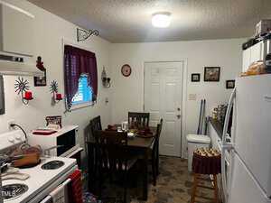 A compact kitchen with white walls features a dining table set for four, a white refrigerator, and an electric stove. Wall decor includes red accents and sunburst designs. A small window with a red curtain is on the left, and the floor has a tiled pattern.