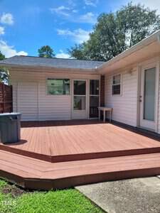 A wooden deck extends from the back of a single-story house with white siding. The deck is empty except for a small table near sliding glass doors. A trash bin sits to the left. The sky is blue with scattered clouds, and trees are visible in the background.