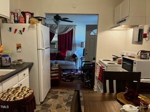 A cozy kitchen and living room are shown. The kitchen has white cabinets, a white fridge, and a small dining table. The living room features a white couch, a red curtain, and a patterned rug. A ceiling fan and a door with a window are visible in the background.