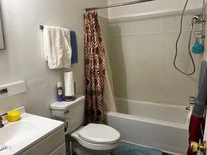 A small bathroom with a white vanity and sink, a toilet, and a bathtub with a colorful shower curtain. Toiletries are placed on the toilet tank. Towels hang on the wall and a blue bath mat is on the floor.