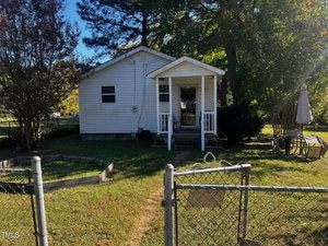 A small white house with a covered front porch is surrounded by trees. The lawn is enclosed by a chain-link fence. An umbrella and chairs are visible in the yard. The sky is clear, and the sun is shining brightly.