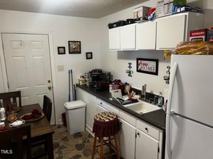 A small kitchen featuring white cabinets, a refrigerator, and a countertop with various appliances and items. A dining table with red napkins is set nearby. Cleaning supplies and a trash bin are visible in the corner. A framed sign decorates the wall.