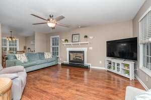 A spacious living room with hardwood floors featuring a gray couch and a recliner. A fireplace with a white mantel is centered against the far wall, and a large flat-screen TV is placed on a stand to the right. A ceiling fan hangs from the ceiling.