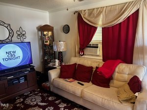 A living room with a beige couch decorated with red pillows and a blanket. An air conditioner is in the window behind red and beige curtains. A TV displaying "New CBS Tonight" is on a stand with a glass display cabinet nearby.