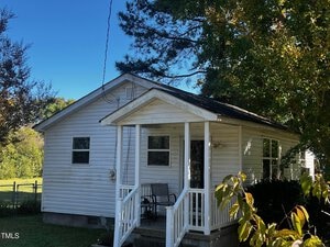A small white house with a porch and two chairs sits amidst a grassy area. Trees surround the house, partially casting shadows on the structure. The sky is clear and blue.
