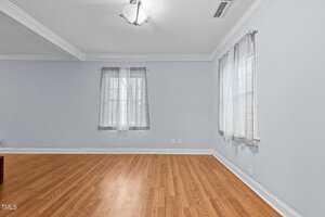 An empty room with light blue walls and wooden flooring. It features two windows with sheer curtains, a white ceiling with crown molding, a modern ceiling light fixture, and two power outlets on one wall.