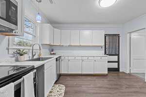 A modern kitchen with white cabinets, a stainless steel oven, and a built-in microwave. There is a potted plant on the countertop, a blue pendant light above, and a window near the sink. The floor is made of wood, and the walls are painted light blue.