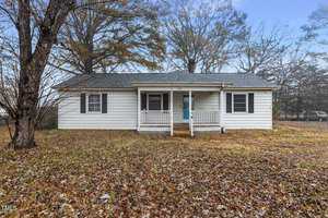 A small, single-story white house at 302 12th Street features a covered porch and two front windows. It is surrounded by bare trees and a lawn blanketed in fallen leaves. The dark gray roof complements the wooden step leading up to the welcoming porch in Butner.