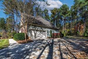 A suburban house with a two-car garage surrounded by trees. The driveway is wide and bordered by shrubs and a deciduous tree. The sky is clear, and there are shadows on the ground from the trees.
