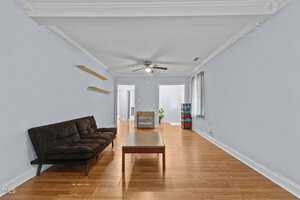 A living room with light blue walls and wooden flooring features a black futon on the left, a wooden coffee table in the center, and wall-mounted shelves. A ceiling fan is overhead, and windows with blinds allow natural light. A doorway is at the far end.