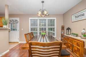 Dining room with a wooden table and six chairs. A checkered runner and a small plant decorate the table. A sideboard with decorative items is against the far wall. Large windows let in natural light. A wall sign reads "home." A chandelier hangs overhead.