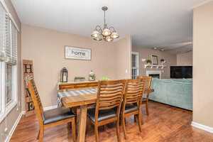 A dining room with a wooden table and six chairs on hardwood flooring. A checkered tablecloth covers the table. A light fixture hangs overhead. The room has beige walls, a wall with framed decor, and an opening to a living area with a green sofa.