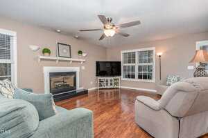 A living room with hardwood floors features a gray couch, a beige recliner, and a fireplace with a white mantel. A large TV sits on a stand nearby. Decorative plants and a framed picture are on the mantel. Ceiling fan and lamp provide light.