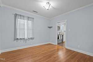 A vacant room with light blue walls, a single window with sheer curtains, and wooden flooring. A doorway leads to a kitchen area with white cabinetry and stainless steel appliances. A modern light fixture hangs from the ceiling.