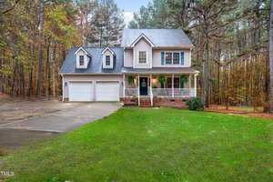 A two-story house with white siding and a gray roof is surrounded by trees. It features a front porch with potted plants, two dormer windows, and a two-car garage. The driveway leads up to the house which is set amid a lush green lawn.