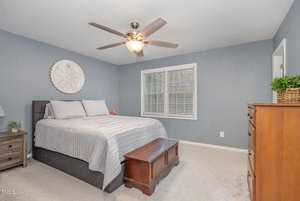 A bedroom with light gray walls, featuring a bed with gray and white bedding. A ceiling fan is overhead, and there are nightstands with lamps on each side. A wooden chest sits at the foot of the bed, and a dresser is against the wall. A window with blinds is visible.