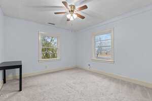 An empty room with light blue walls and beige carpet. Two windows provide natural light, and a ceiling fan with wooden blades is centered on the ceiling. A small black table is in the corner near one window.