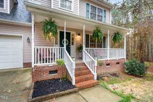 A charming two-story house at 4119 Tall Pine Drive, nestled in Franklinton, boasts beige siding and a covered porch adorned with potted ferns. The white railings and red brick steps add classic elegance, while a small shrub enhances its corner. Surrounded by trees, it features a convenient paved driveway.