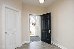 An open black front door with a silver knob leads from a beige-walled room with light wood flooring to a driveway outside. A white door is on the left, and bare trees can be seen through the doorway.