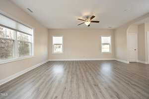 Spacious empty room with light wood flooring and beige walls. Two large windows bring in natural light. A ceiling fan is centered on the ceiling. Doorways lead to adjoining rooms.
