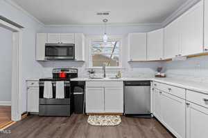 A modern kitchen with white cabinets and marble countertops. There is a stainless steel oven with a microwave above it, a dishwasher, and a window with a view of trees. A pendant light hangs from the ceiling, and a small indoor plant is on the counter.