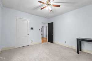 An empty room with light blue walls and beige carpet. A ceiling fan with wooden blades is centered above. The room has a white door and a black door, with a narrow black table against one wall. A section of hardwood floor is visible through the doorway.