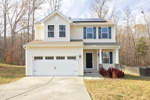 Nestled on 111 Richmond Run, this two-story beige house features a double garage and solar panels on the roof, next to green shutters. Surrounded by a lightly wooded area with autumn foliage and a concrete driveway leading to a small front lawn, it's an ideal setting for sustainable living.