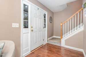 Entrance hallway featuring a white front door with glass panels, beige walls, and a wooden stairway with white railings. The floor is hardwood, and a light switch is visible on the wall. A decorative wreath hangs on the wall by the stairs.