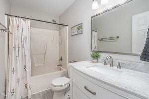 A clean, modern bathroom with a white toilet, a white shower curtain adorned with floral patterns, a bathtub, a large mirror above a marble-topped vanity, and a potted plant. The floor is wood-patterned, and there are two wall-mounted lights.