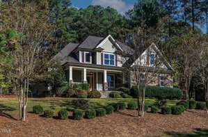 Nestled at 3580 Garner Terrace Way in Wake Forest, a charming two-story house with a gabled roof sits behind a manicured lawn and neatly trimmed shrubs. The exterior boasts stone and siding, while the porch is supported by columns. In the background, tall trees reach towards the partly cloudy sky.