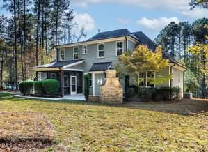 A two-story gray house with white trim sits amidst tall trees. The front yard has a grassy area and shrubs. There's a stone chimney on the side and a screened porch on the left. The sky is partly cloudy, and sunlight filters through the trees.