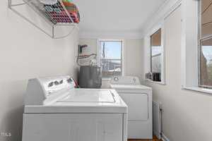 Laundry room with a white washing machine and dryer side by side against a pale wall. Above them is a wire shelf holding colorful towels. The room has a window to the right and a larger window ahead, both letting in natural light.