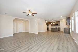 A spacious, open-concept living area with light wood flooring. It features a ceiling fan, recessed lighting, and a view into a modern kitchen with an island and pendant lights. White walls and large windows provide natural light.