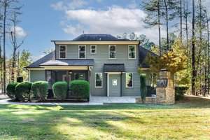 A two-story house with gray siding, featuring a front porch and multiple windows. The surrounding area has neatly trimmed bushes, a stone pillar chimney on the right, and a well-kept lawn with tall trees in the background under a partly cloudy sky.