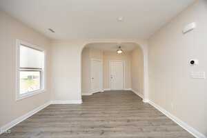 A room with light gray hardwood flooring and beige walls. A large archway leads to a hallway with a white door. There is a window on the left and a small ceiling light fixture. A thermostat is on the right wall.